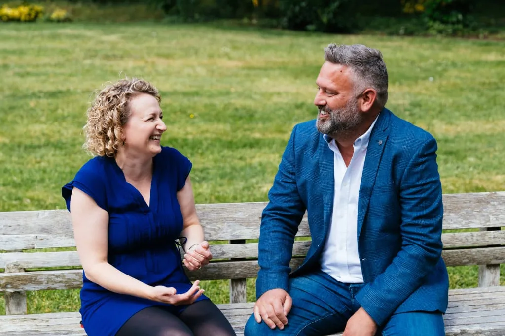 Middle Aged Man and Woman Sat on Park Bench Talking Mobile