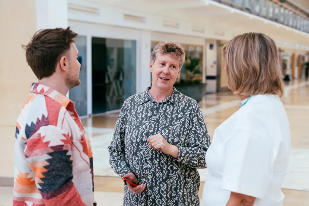 Middle Aged Woman With Short Hair and Patterned Shirt Stood Talking to Two Colleagues Mobile