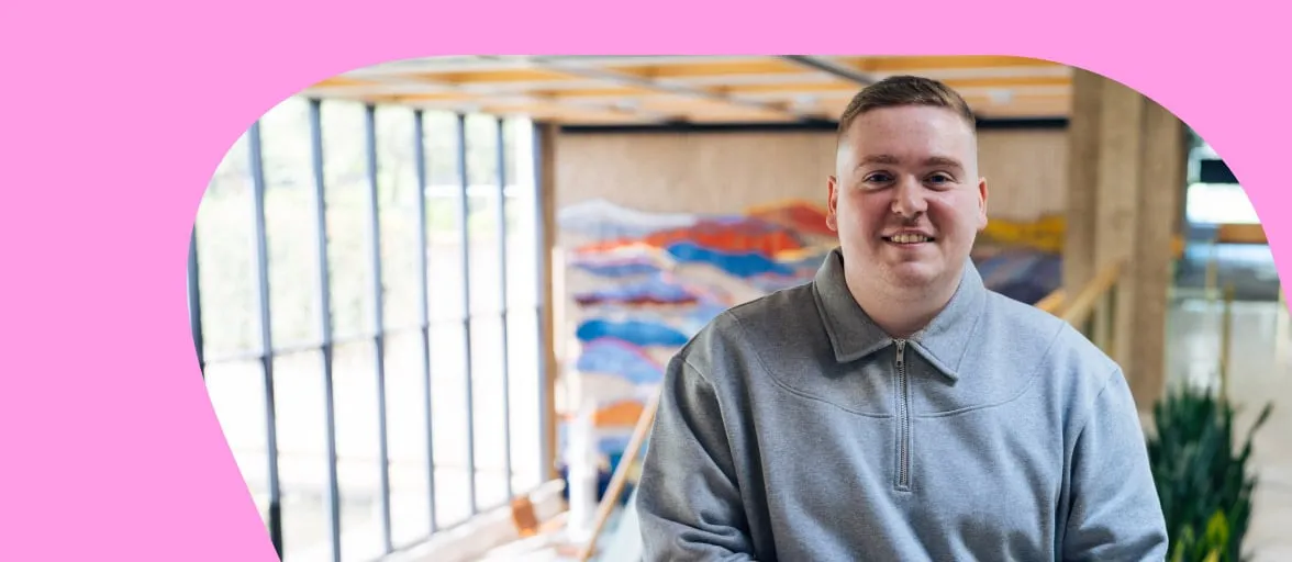 Young Man Wearing Grey Quarter Zip Sweater Stood in Soft Focus Colourful Modern Office 1