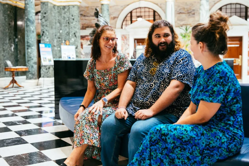 Two Women and Man Wearing Bright Patterned Clothing Sat Talking Mobile