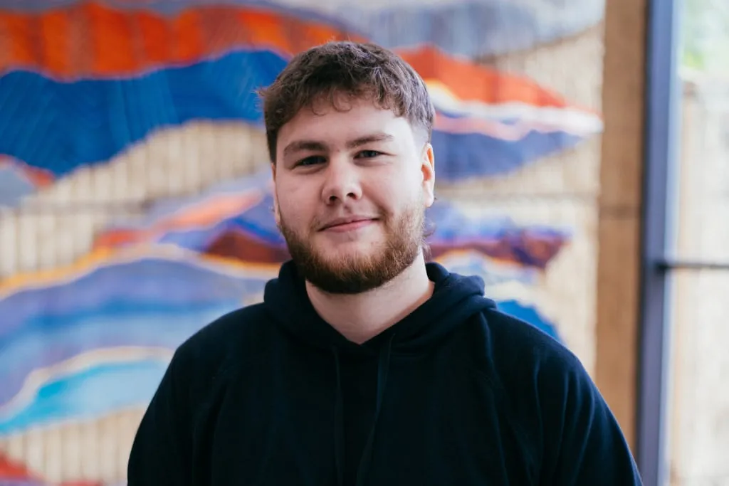 Young Man With Beard and Mullet Haircut Wearing Dark Hoody Against Colourful Soft Focus Background Mobile